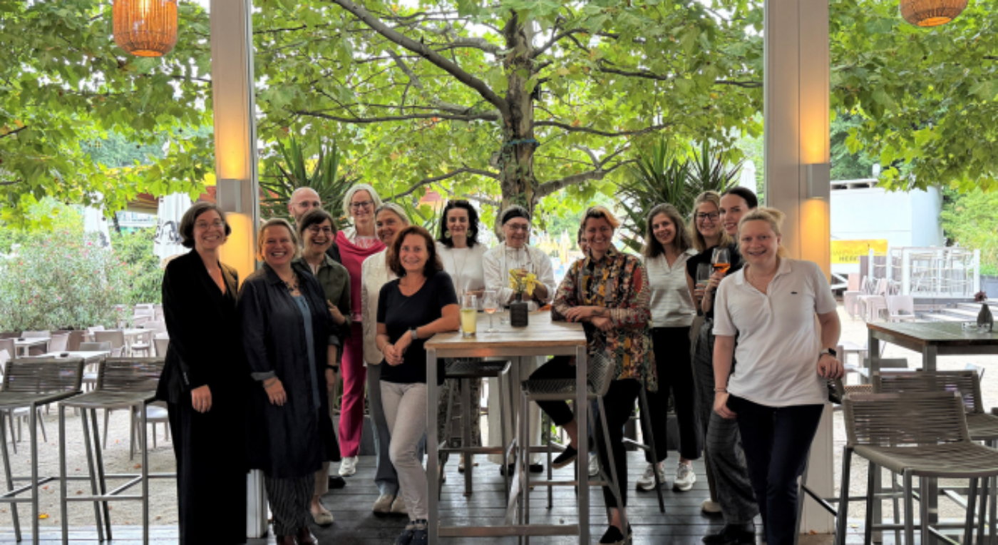 Eine Gruppe Frauen steht für ein Gruppenfoto zusammen. Im Hintergrund sind Stühle und Tische.