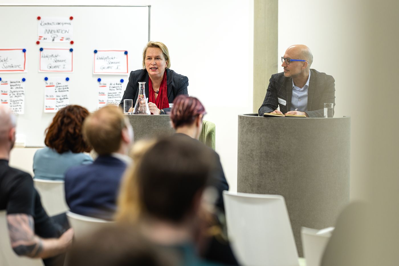 Eine Frau und ein Mann sitzen an einem Podium. Im Vordergrund sieht man das Publikum von hinten.
