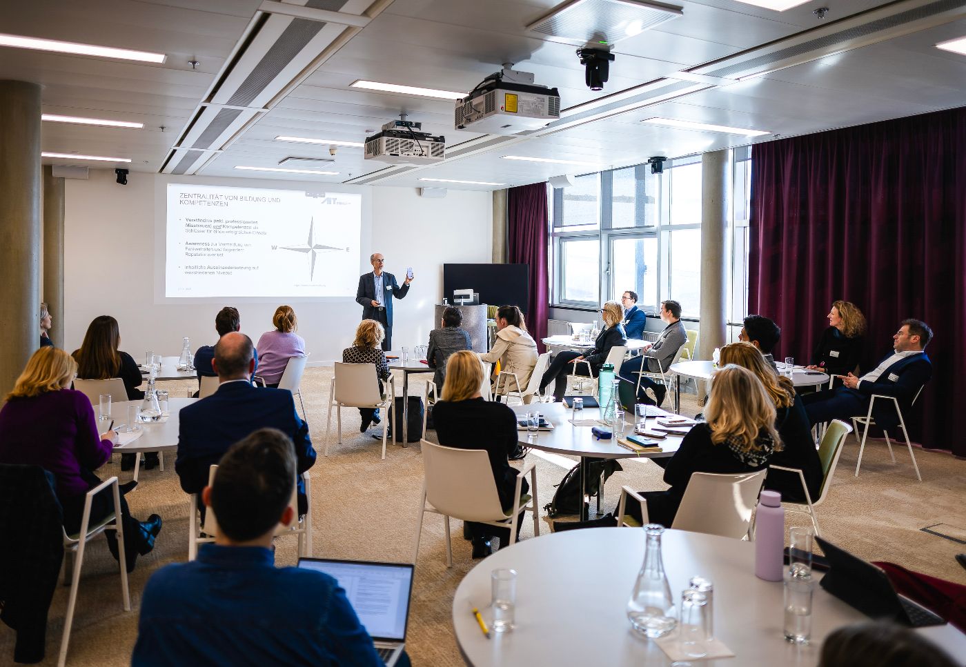 Vortragender im Seminarraum mit Teilnehmenden die an runden Tischen sitzen.
