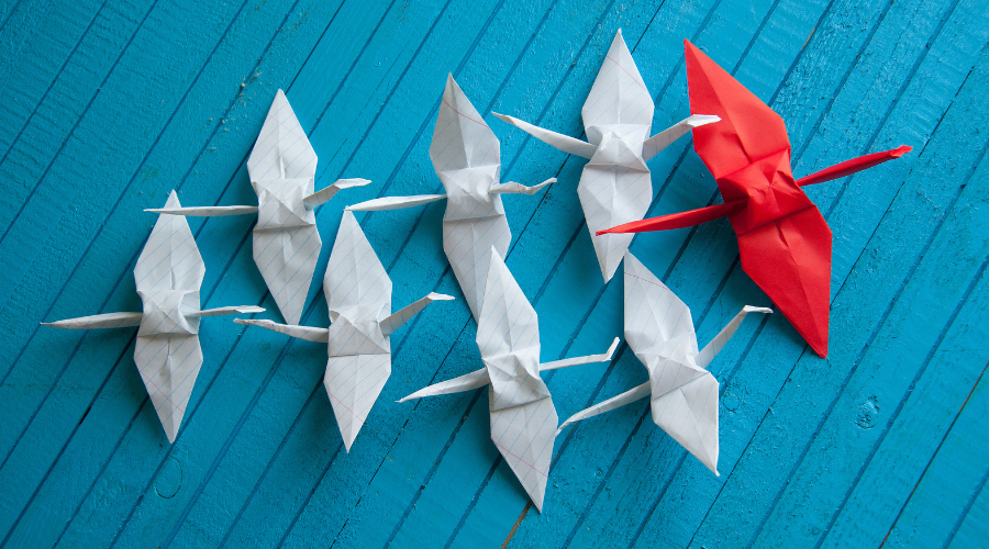 Weiße Origami Kraniche folgen einem roten Origami Kranich. Der Hintergrund ist blau.