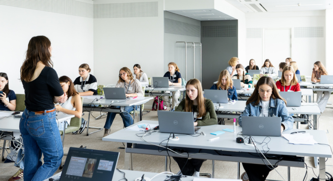 Schülerinnen sitzen vor Computern. Eine Vortragende steht auf der rechten Seite.