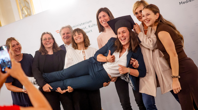 Sechs Teilnehmerinnen des Gender Trainee Programmes halten eine Kollegin liegend in die Höhe. Die gehaltene Frau trägt einen Akademikerhut und gibt einen Daumen nach oben.