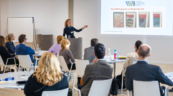 Abteilungsleiterin Petra Dietrich zeigt auf eine Präsentation. Die Zusehenden sieht man von hinten an runden Tischen sitzen.