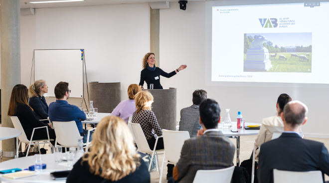 Eine Vortragende zeigt auf ihre Präsentation. Im Vordergrund sieht man die Teilnehmenden von hinten an Tischen sitzen.