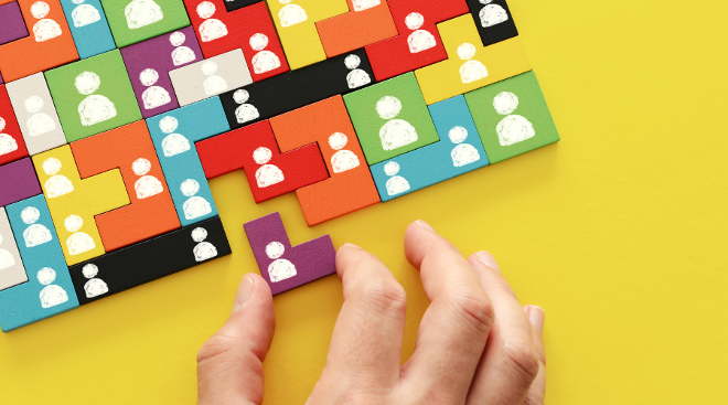 Ein gelber Hintergrund auf dem bunte Holzbausteine wie ein Puzzle liegen. Auf den Bausteinen sind weiße Spielfiguren gemalt. Eine Hand schiebt einen lila Baustein in eine passende Lücke.